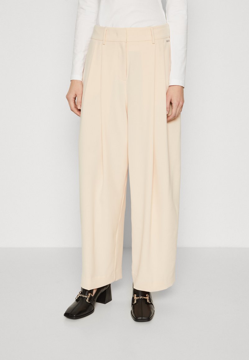 Helle beigefarbene weite Hose mit Falten, mit einer glatten Textur und Gürtelschlaufen. Kombiniert mit schwarzen Blockabsatzschuhen mit goldenen Akzenten.