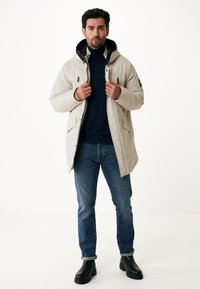 Světle béžová parka s kapucí, se zipovým zapínáním, dvěma bočními kapsami a texturovanou tkaninou. Kombinováno s tmavými džíny a černými botami.