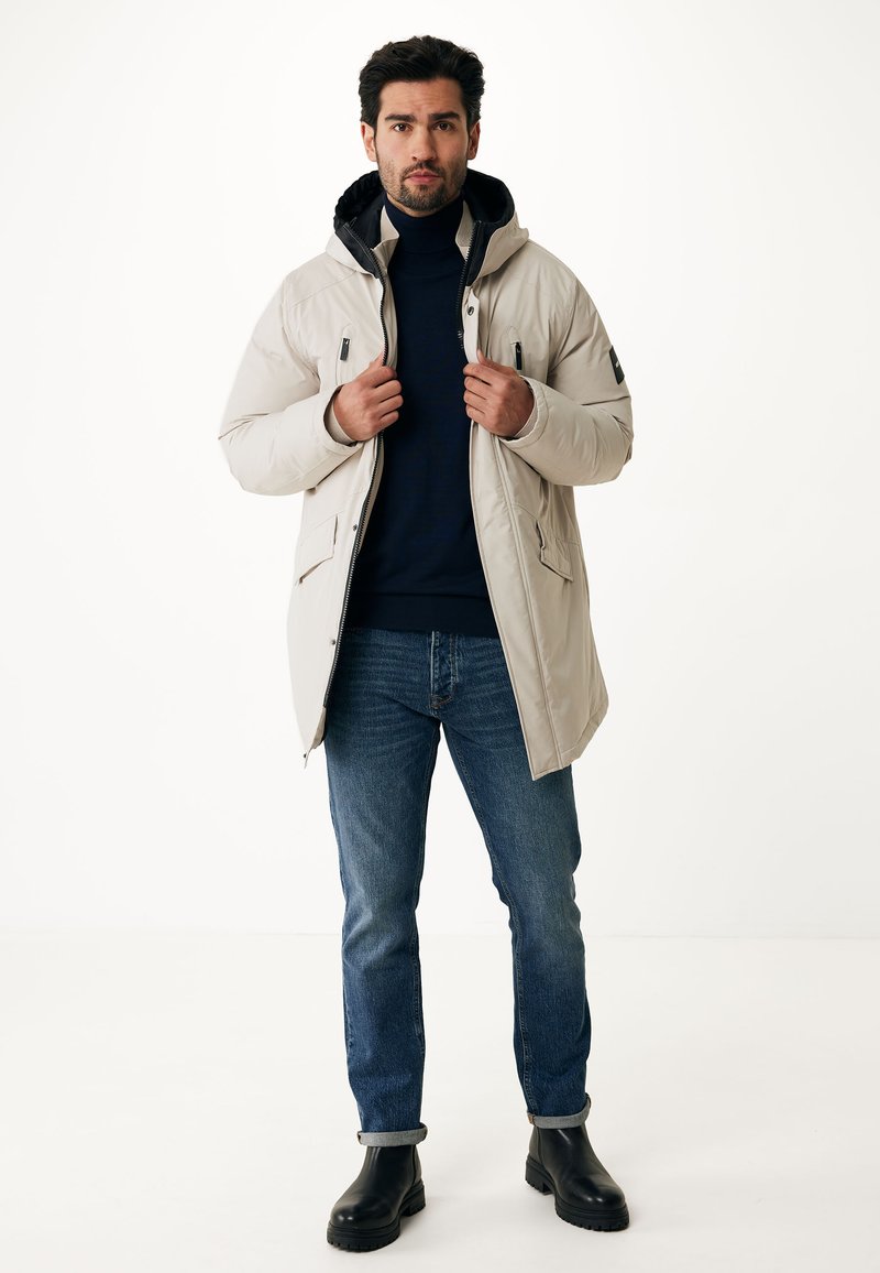 Parka de color beige claro con capucha, que cuenta con cierre de cremallera, dos bolsillos laterales y una tela texturizada. Combinada con jeans oscuros y botas negras.