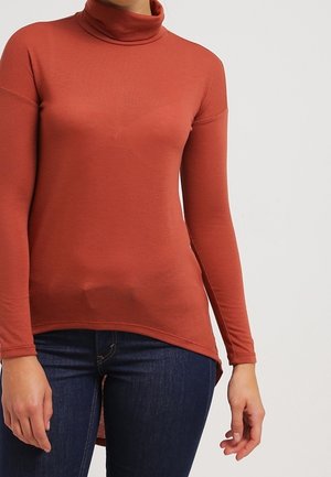 Ruskikansinen turtleneck-toppi, jossa on pitkät hihat, tiukka siluetti ja pyöristetty helma, yhdistettynä tummansinisiin farkkuihin. Sileä kankaan pinta.