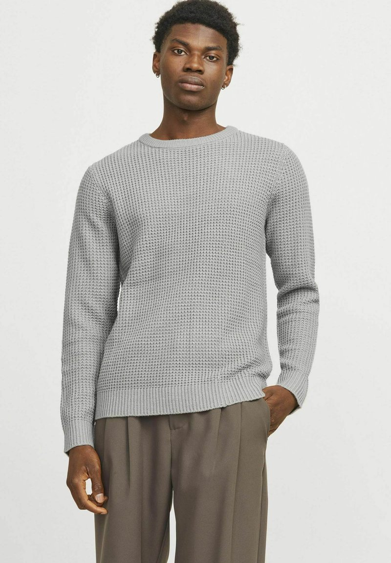 Jersey de punto gris con un patrón texturizado, cuello redondo, puños y dobladillo acanalados, usado sobre pantalones beige. Diseño sencillo y moderno.
