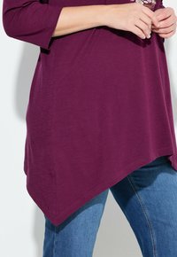 Kurzarmpullover in tiefpurpur mit abgerundetem Saum, weichem Griff und floralen Verzierungen; kombiniert mit blauen Jeans.