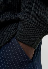 Pull noir en tricot épais avec des poignets côtelés associé à un pantalon à rayures bleu marine. Contraste de texture bien visible entre les matériaux.