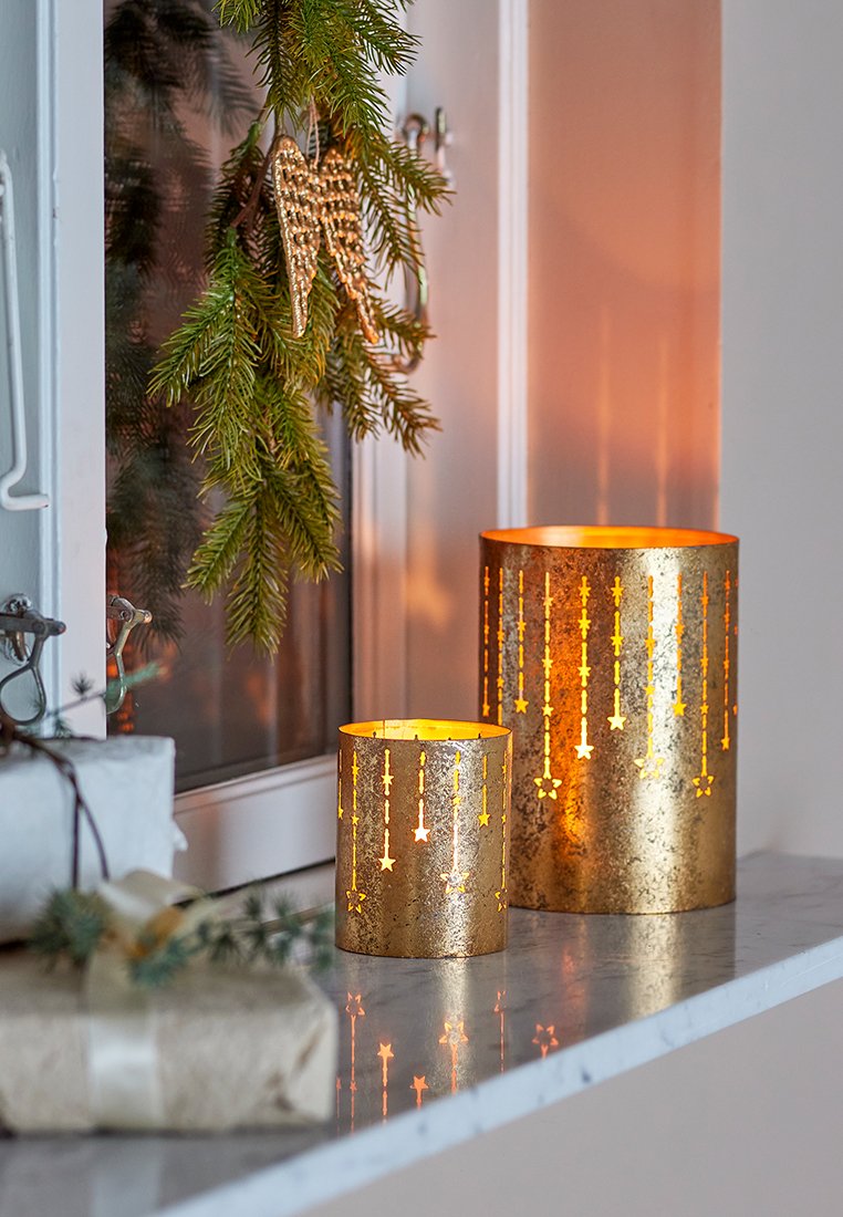Goldene Kerzenhalter aus Metall mit durchbrochenen Sternmustern, die ein warmes Licht abgeben. Ein Halter ist höher, der andere niedriger, und sie stehen auf einer weißen Oberfläche.