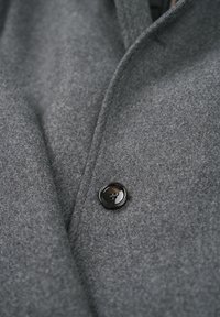 Grauer Wollmantel mit einem schwarzen Knopf mit vier Löchern, strukturiertem Stoff und einem Reverskragen-Design. Nahaufnahme, die die Details betont.