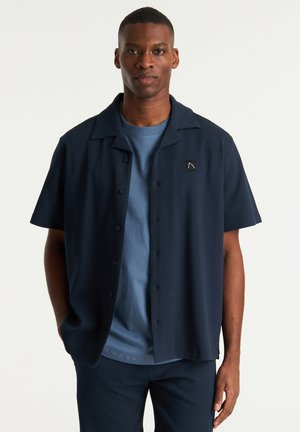 Marineblaue Kurzarmbluse mit einem strukturierten Muster, Knopfschluss und einem dezenten Logo-Patch, getragen über einem hellblauen T-Shirt.