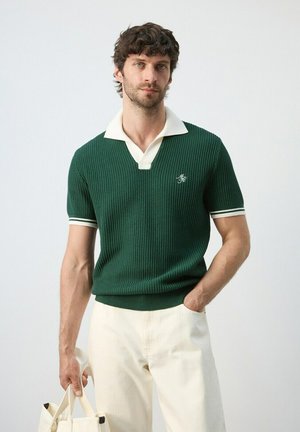 Hombre con camiseta polo de manga corta texturizada verde, pantalones crema, sosteniendo un bolso tote beige, de pie contra un fondo claro liso.