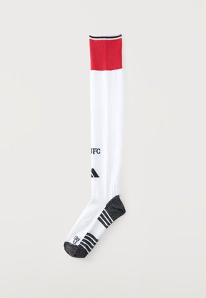 Chaussette montante avec une bande rouge en haut, jambe blanche, pied et bout noirs, et détails du logo noirs près de la cheville et du tibia.