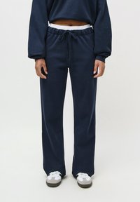 Persona che indossa pantaloni della tuta blu navy con cinturino elastico bianco e stringhe, abbinati a sneaker bianche e grigie su sfondo bianco.