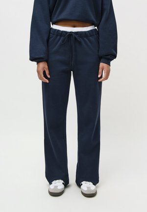 Person wearing navy blue sweatpants with white elastic waistband and drawstring, paired with white and gray sneakers on white background.