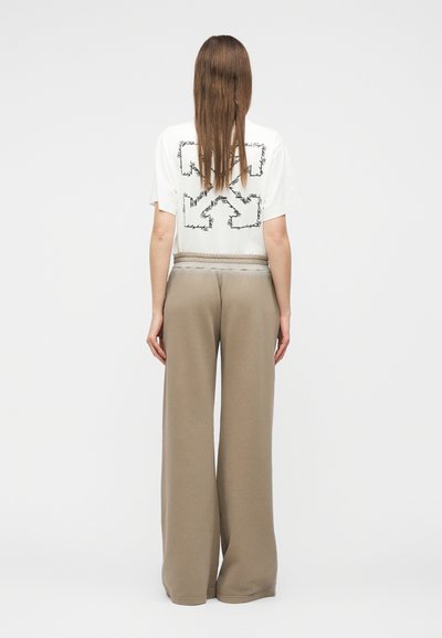 Γυναίκα με μακριά ίσια μαλλιά, που φοράει λευκό T-shirt με μαύρα αφηρημένα βέλη και φαρδιά μπεζ παντελόνια, στέκεται μπροστά σε απλό λευκό φόντο.