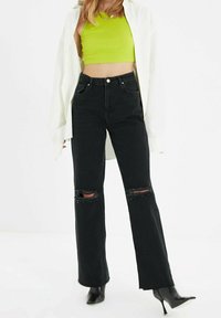 Jeans neri a vita alta con strappi sulle ginocchia, abbinati a un crop top verde brillante e a una camicia bianca oversize, che mettono in risalto un look casual.