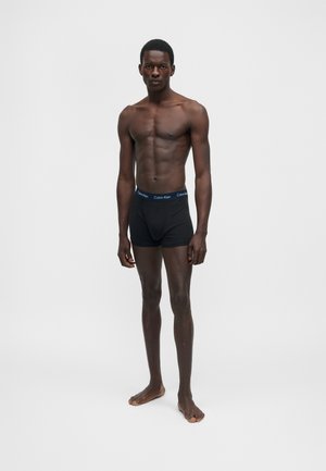 Λεπτός άνδρας με σκούρο δέρμα, όρθιος ξυπόλυτος, φοράει μαύρα εσώρουχα Calvin Klein boxer μπροστά σε απλό φωτεινό φόντο.