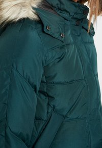Veste d'hiver matelassée vert foncé avec capuche bordée de fourrure et boutons-pression au col, portée par une personne partiellement visible du cou jusqu'en bas.