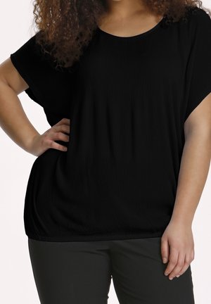 Woman wearing a loose black short-sleeve top and black pants, standing with one hand on hip and curly hair partially visible.