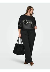 Femme portant un pull noir avec le texte "love" et des rayures, un pantalon noir, des chaussures marron, portant un grand sac fourre-tout noir, debout sur fond blanc.