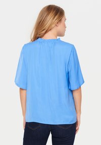 Blusa blu a maniche corte con un sottile motivo a pois e un colletto arricciato. Il tessuto appare leggero e liscio. Abbinata a jeans scuri.