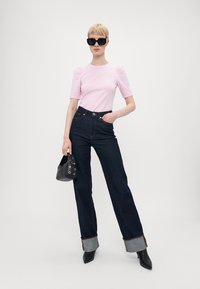 Rozā pieguļošs tops ar puffed piedurknēm, augsta josta tumšās džinsās ar kontrastējošu vīlējumu, ar elastīgām pēdām, melna rokassoma un eleganti melni zābaki.