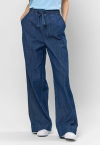 Pantaloni largi din denim închis, cu o talie elastică și șnur, buzunare laterale și o textură fină. Purtați cu adidași albi.