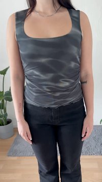 Persona che indossa un top satinato senza maniche grigio scuro e pantaloni neri, in piedi all'interno con piante in vaso e un tappeto sullo sfondo.