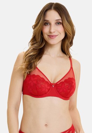 Donna con capelli lunghi e mossi che indossa un reggiseno rosso in pizzo con ferretto, sorridente davanti a uno sfondo chiaro e uniforme.