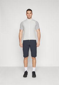 Maillot de cyclisme gris clair à manches courtes avec fermeture éclair à l'avant, associé à un short bleu foncé avec des détails de logo blancs. Porté avec des chaussures noires et des chaussettes blanches.
