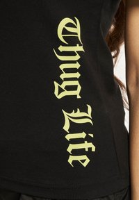 Černý bavlněný tílko s velkým žlutým gotickým písmem "Thug Lite" vertikálně podél strany. Jednoduchý design, ribovaná textura.