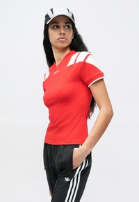 Camiseta deportiva roja con rayas blancas en los hombros, mangas cortas y diseño de cuello en v. Combinada con pantalones deportivos negros con tres rayas blancas.