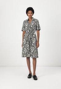Abito camicia floreale in bianco e nero con maniche corte, vita aderente e gonna fino al ginocchio. Completato con un colletto e dettaglio con fiocco.