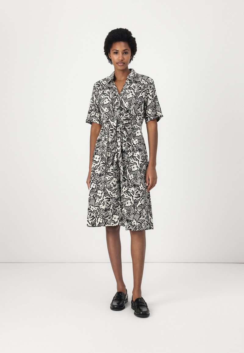 Abito camicia floreale in bianco e nero con maniche corte, vita aderente e gonna fino al ginocchio. Completato con un colletto e dettaglio con fiocco.