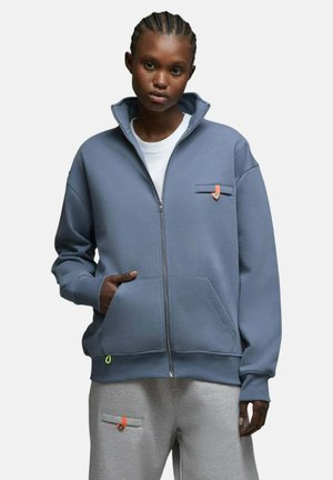 Jeune personne portant une veste bleue zippée et un pantalon de survêtement gris avec des détails de boutons orange, debout avec une main dans la poche.