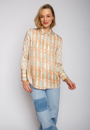 Cremfarbendes Hemd mit Knöpfen und geometrischem Muster in Pfirsich, Blau und Beige; kombiniert mit hellblauen, weiten Jeans mit Patches.