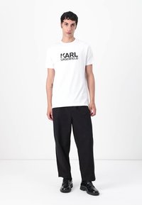 Hvit bomull t-skjorte med en svart "KARL LAGERFELD" logo, sammen med løstsittende svarte bukser og svarte sko.