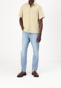 Beige shirt met korte mouwen en een zak, gecombineerd met lichtblauwe jeans en bruine instappers. Het shirt heeft een knoopsluiting aan de voorkant en een gladde textuur.
