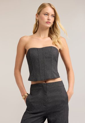 Mujer rubia que lleva un top gris oscuro ajustado sin tirantes y pantalones a juego de talle alto con pendientes de aro dorados y pulseras.