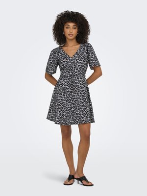 Femme aux cheveux bouclés debout, vêtue d'une robe à manches courtes imprimé léopard noir et gris, et de sandales à talons noires sur un fond gris.