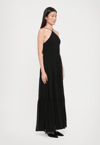Svart maxi-kjole med en tettsittende, smocket bodice og en flytende, lagdelt skjørt. Har tynne, kryssede stropper og myk, lett stoff.