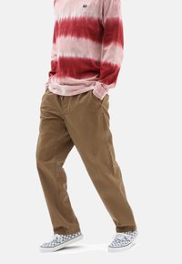 Pantalon marron ample avec des poches latérales, associé à un t-shirt à manches longues tie-dye rouge et blanc et des chaussures à enfiler à carreaux.