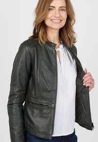 Mörkgrön läderjacka med rund halsringning, dragkedja fram och stickade detaljer på axlarna, i kombination med en vit blus och denimjeans.