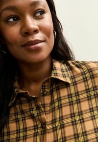 Camisa de cuadros en tonos marrón y amarillo, con cuello abotonado, tela texturizada y vista en primer plano del escote y el rostro.