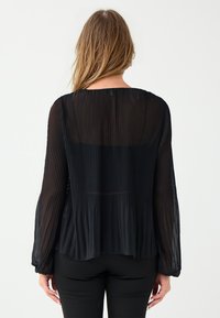Schwarze, plissierte Bluse mit langen Ärmeln und lockerer Passform, ausgestattet mit einem hohen Kragen und fließendem Stoff, der gut zu enganliegenden schwarzen Hosen passt.