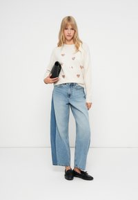 Beżowy sweter z metalicznymi wzorami serca, połączony z jasnoniebieskimi spodniami o szerokich nogawkach z denimu, oraz czarną torebką kopertową. Czarne buty.