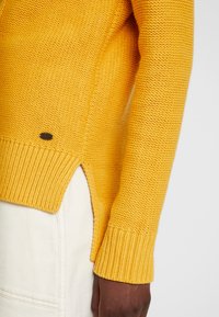 Gestrickter gelber Pullover mit gerippten Bündchen und Saum, seitlichen Schlitzen und einem kleinen Logodetail am Saum. Weiche Textur und mittleres Gewicht.