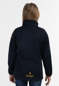 Sudadera de forro polar azul marino con cuello alto, variaciones en la textura y detalle del logo amarillo en el dobladillo trasero. Mangas largas.