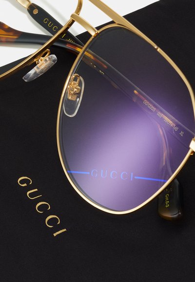 Gold-framed sunglasses with gradient purple lenses, showcasing a 'Gucci' logo. Accompanied by a black case with gold lettering.