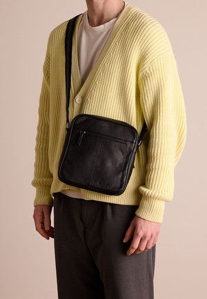 Zwart leren crossbody tas met een gestructureerd oppervlak, vierkante vorm, een voorzak met rits en een verstelbare schouderriem, gedragen over een lichtgele cardigan.