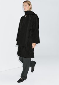 Black oversized coat with a high collar, paired with dark grey wide-leg trousers and black slip-on shoes. Smooth fabric texture throughout.