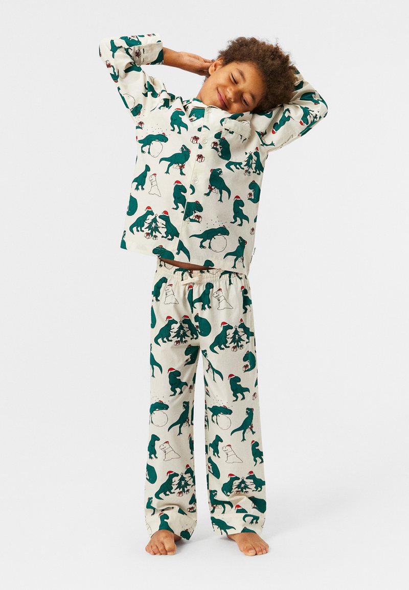 Pidžama set s krem pozadinom i uzorcima zelenih dinosaura i blagdanskim motivima. Košulja s gumbima, hlače s elastičnom gumom.