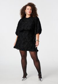 Zwart jurk met gemêleerd bloemenborduurwerk, pofmouwen en een getailleerde taille. Gecombineerd met glanzende zwarte kousen en zilveren hakken.