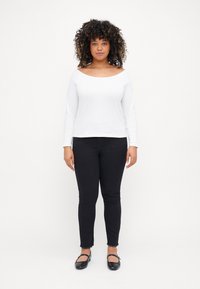Camisa blanca de manga larga con un diseño off-shoulder, combinada con jeans ajustados negros y zapatos planos negros. Los jeans tienen dobladillos deshilachados.